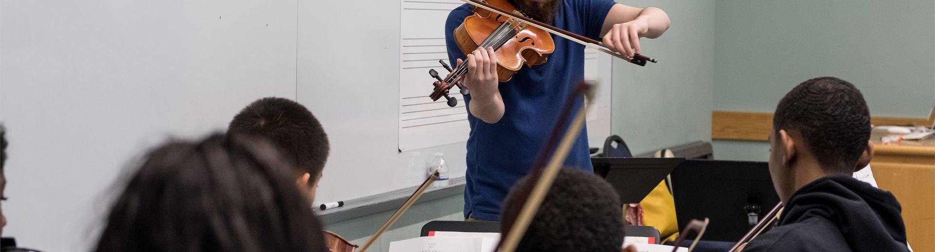 A violin teacher plays at the front of the class in front of young students.